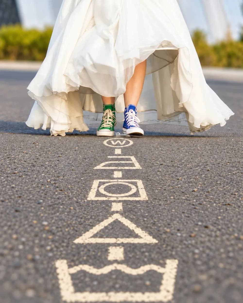 Une mariée en robe blanche et baskets montantes avance sur une chaussée où sont peints les symboles d’entretien textile : aquanettoyage, séchage, repassage, blanchiment et lavage